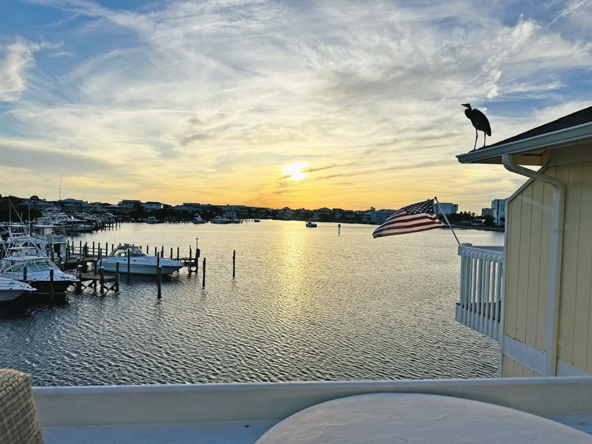 The hero image captures a serene harbor at sunset, with boats docked along the water, a gentle pastel sky, an American flag fluttering atop a building, and a heron perched on the roof—evoking a peaceful, coastal charm.