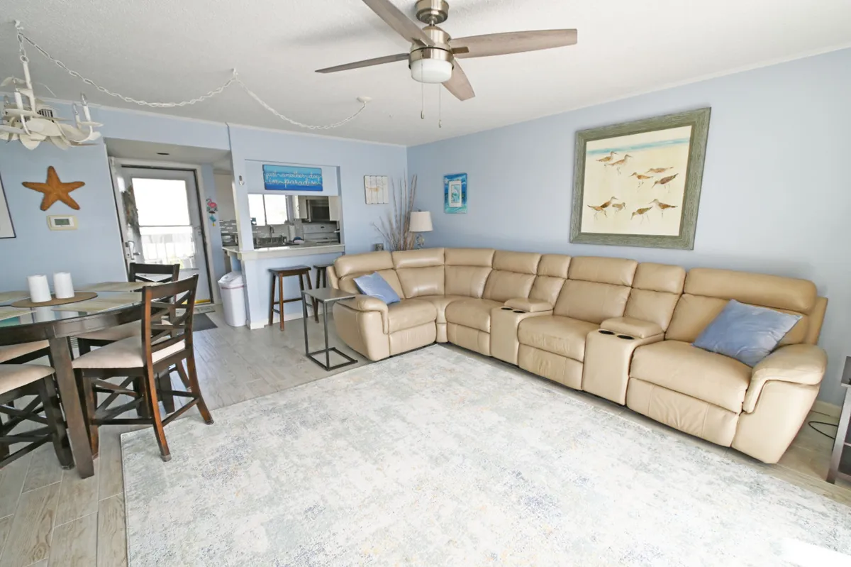 This image presents a spacious open-concept living area featuring a large beige sectional sofa with recliners, a round dining table with chairs, coastal-themed wall art, and a kitchen pass-through window with bar stools. The space is bright, airy, and designed for both comfort and socializing.