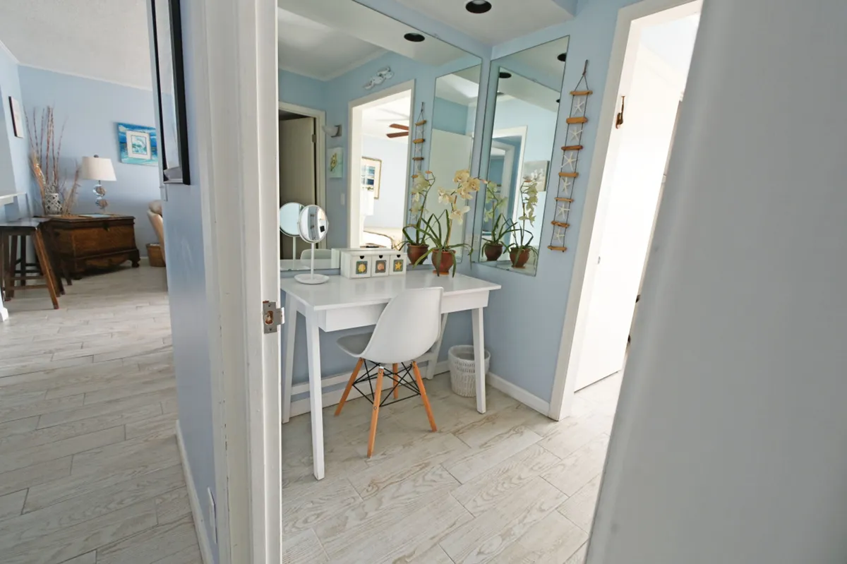 A stylish white vanity desk with mirror and orchid plants is paired with a modern white chair. Large mirrors reflect soft blue wall color, enhancing the sense of space and tranquility. The setup is ideal for makeup application or as a functional workspace, while the decor suggests moments of calm before social activities.