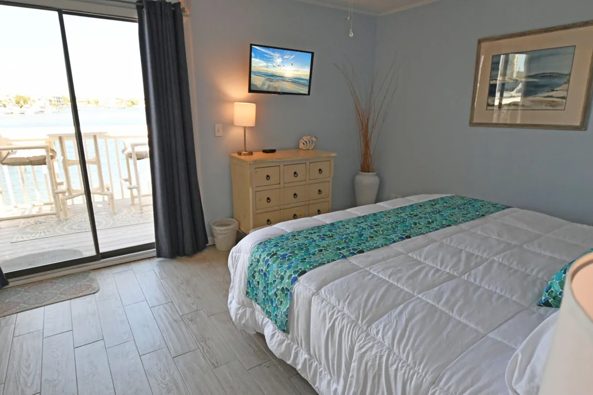 A king-sized bed with a decorative runner and plush pillows sits across from a wall-mounted flat-screen TV displaying a serene beach scene. A sliding glass door opens directly to a balcony with a harbor view, and a light wooden dresser with a lamp and decorative shell adds a touch of coastal charm.