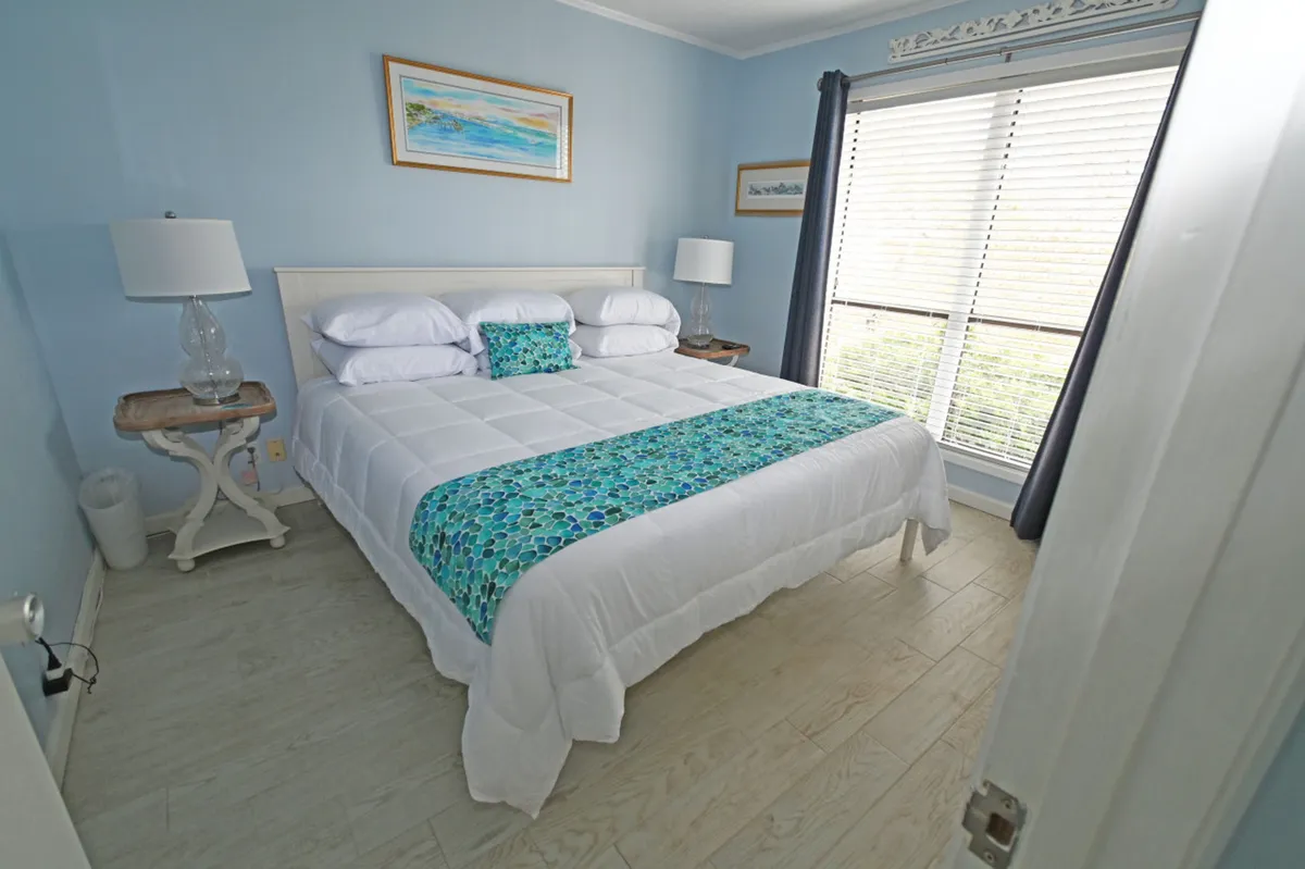 A peaceful bedroom featuring a king-sized bed with white bedding and a blue decorative runner. The room is brightened by a large window with blinds and dark curtains, and matching bedside tables with glass lamps add symmetry. Coastal-themed wall art completes the serene ambiance.