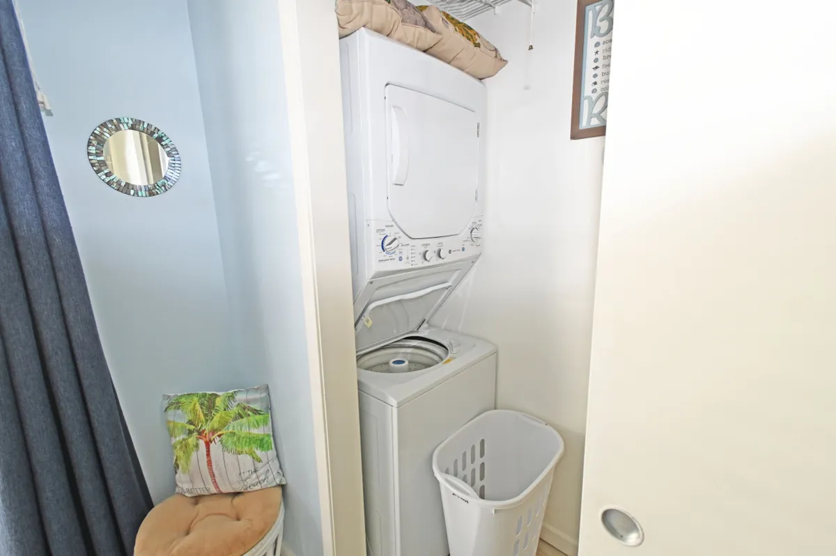 A cozy space featuring a stacked washer and dryer unit tucked into a corner, alongside a laundry basket and a cushioned bench with a decorative palm tree pillow. The area exudes practical charm with a homey feel, enhanced by a decorative mirror on the wall.