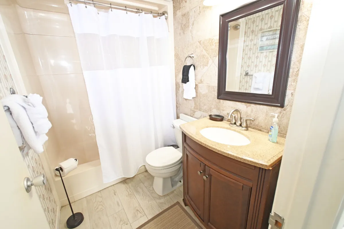 A clean, modern bathroom featuring a wooden vanity with granite countertop and sink, a white shower curtain enclosing a bathtub, a toilet, and a large framed mirror above the vanity. The mood is calm and refreshing, suggesting a well-appointed space for grooming and relaxation.