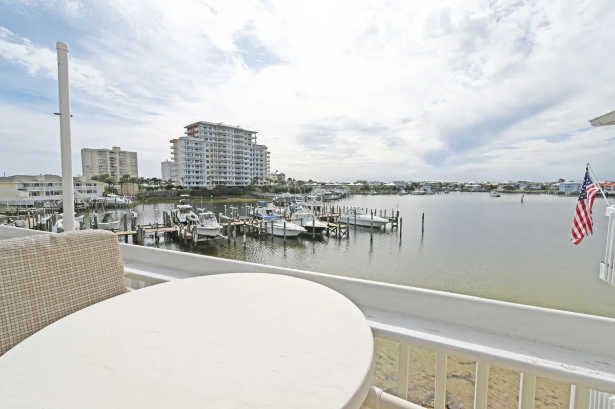 A round outdoor table with a cushioned chair sits on a balcony overlooking a bustling waterfront marina dotted with docked boats. In the background, condominium buildings and an American flag on the railing set the scene for relaxed mornings or evenings spent watching harbor activity.