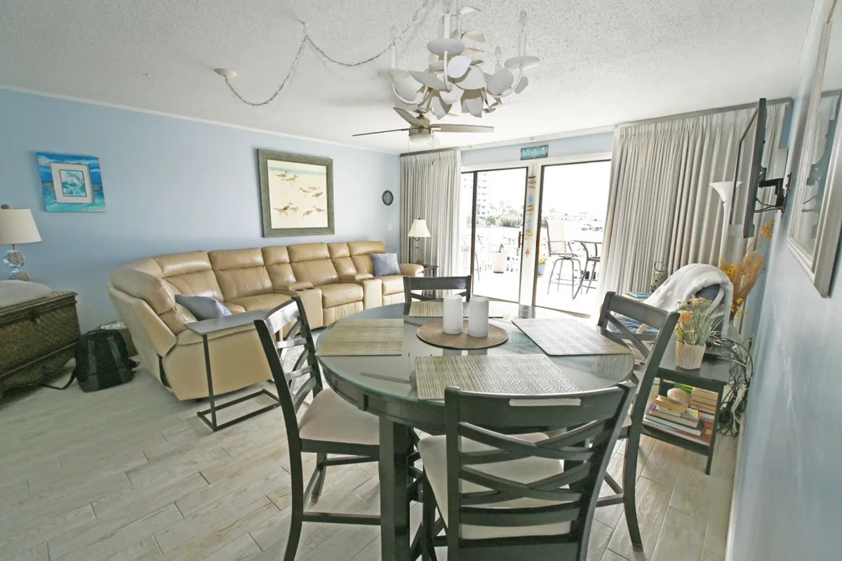 A spacious and comfortable living area with a round glass dining table, a large beige leather sectional sofa, and sliding glass doors leading to an outdoor balcony. The room is bathed in natural light and offers a welcoming space for family movie nights or board games after a day of adventure.