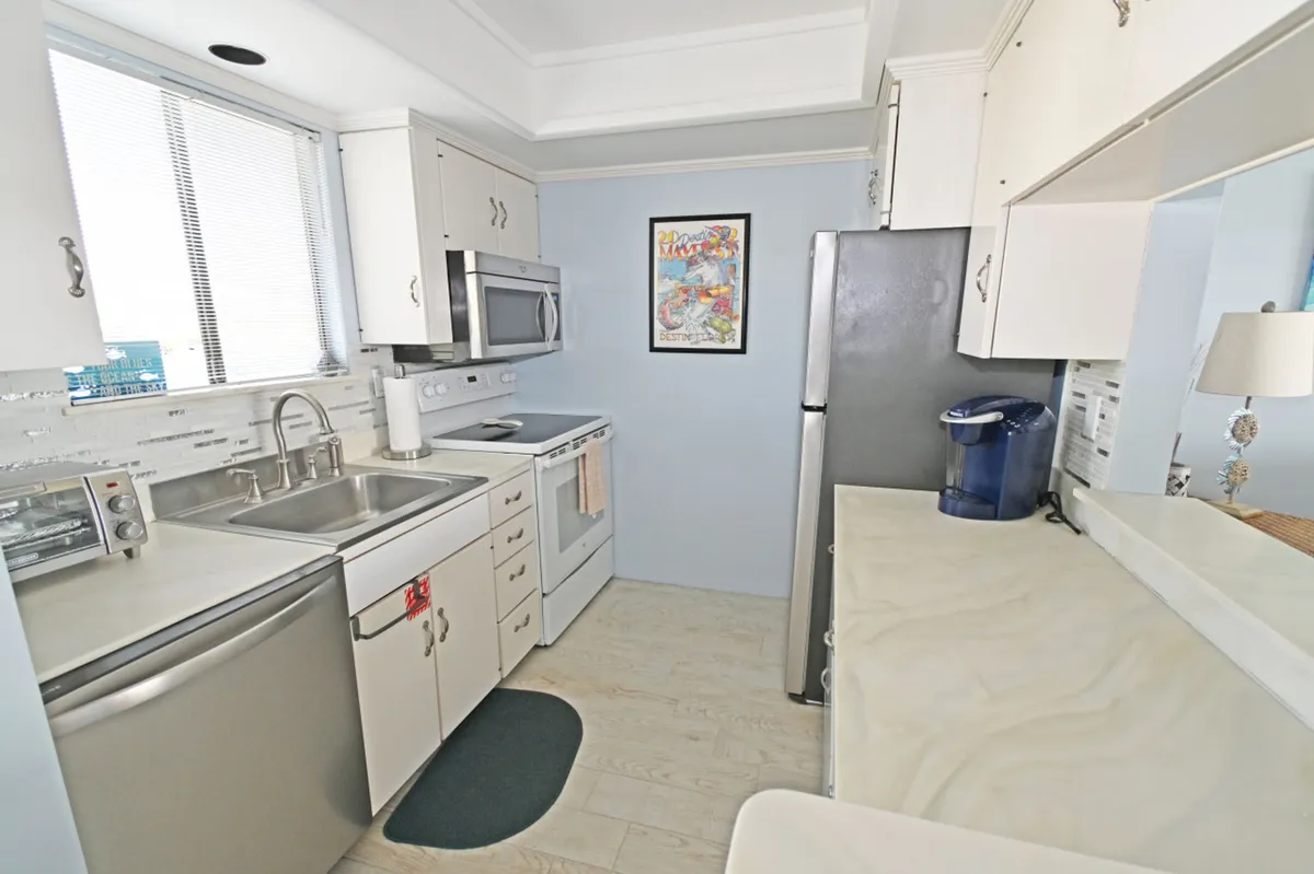 A bright, well-equipped kitchen with stainless steel appliances including a refrigerator, microwave, dishwasher, and stove. A double sink sits under a window, and a coffee maker is visible on the counter. The kitchen is decorated with cheerful coastal artwork.