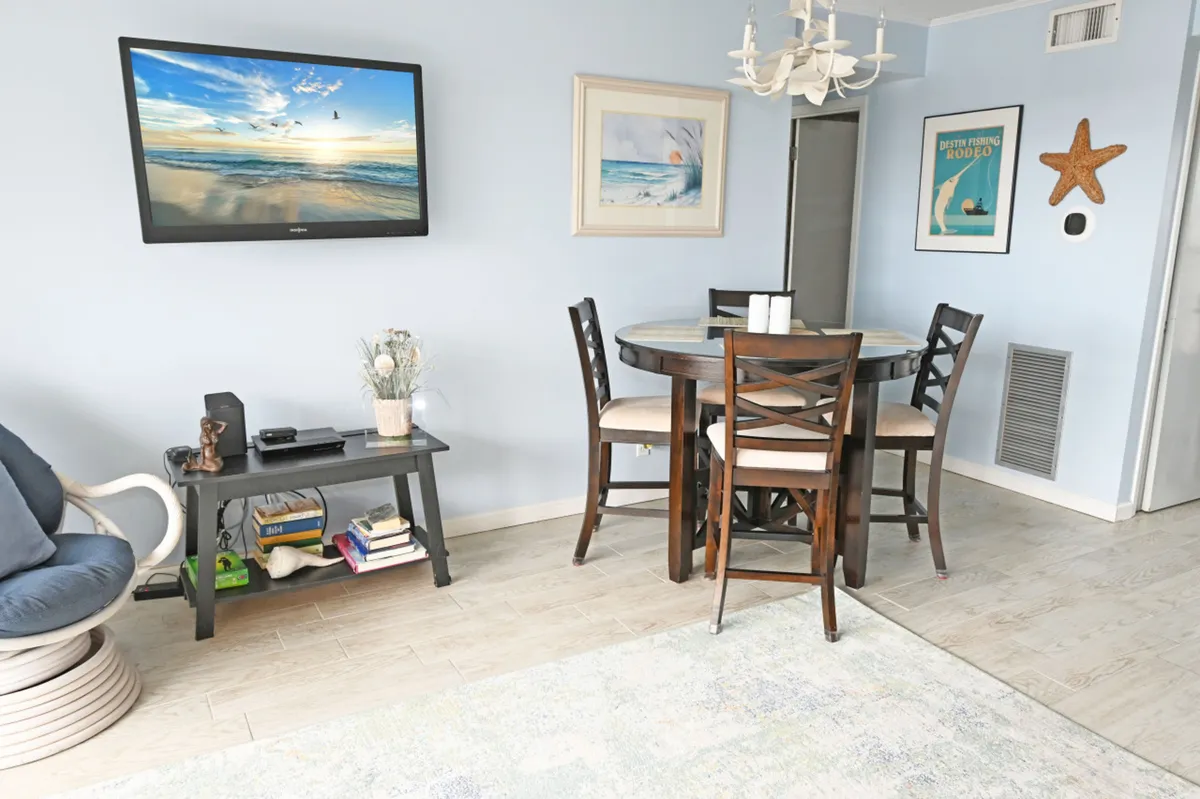 A bright, inviting dining area with a round table and four chairs, a comfortable swivel chair, and a wall-mounted TV displaying a coastal scene. The space is decorated with beach-themed art and offers a relaxed, sociable atmosphere ideal for unwinding after active days.