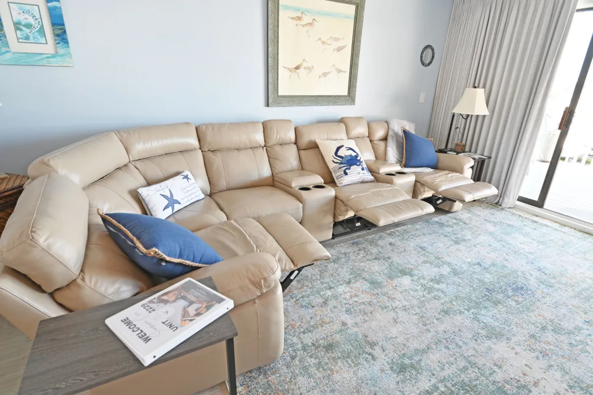 A cozy living area with a beige sectional recliner sofa adorned with coastal-themed pillows, a sliding glass door opening to a private deck, and stylish coastal artwork. The space is designed for relaxation, with direct indoor-outdoor flow to the harborview deck.