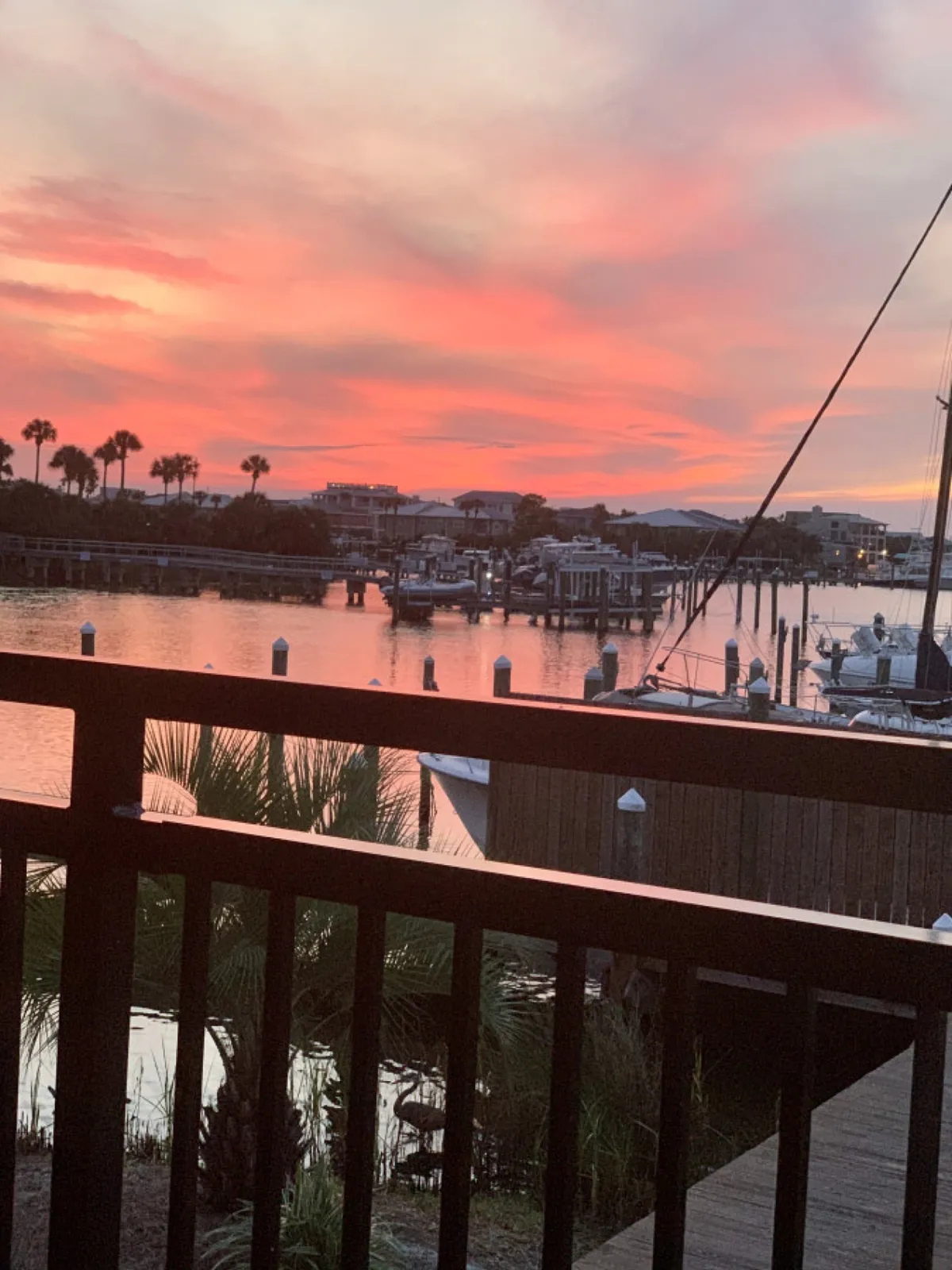 A dramatic sunset sky with pink and orange hues stretches over a tranquil harbor filled with boats and docks, framed by silhouetted palm trees and a wooden balcony railing in the foreground. The image evokes a sense of peacefulness and awe as the day transitions into evening.