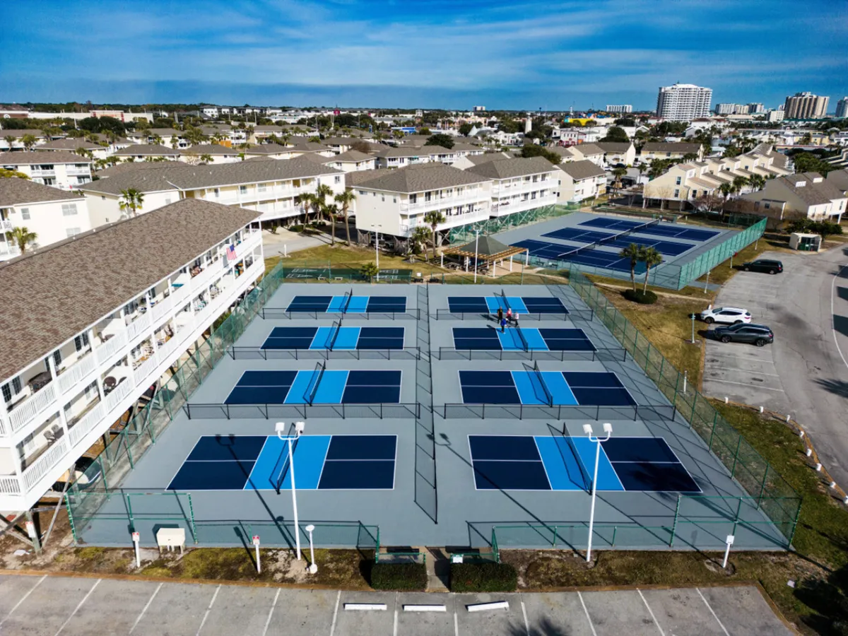 A lively exterior scene showing multiple pickleball courts in action, set against the backdrop of resort buildings. Players are engaged in a match, with several onlookers nearby, capturing the energetic and social spirit of resort amenities.