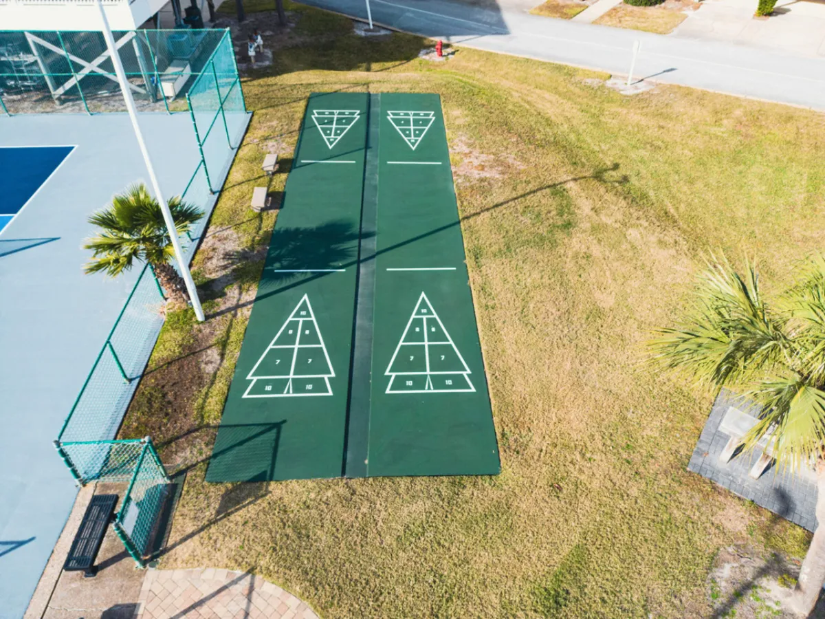 A close-up exterior shot of shuffleboard courts with clear markings, a nearby tennis or pickleball court, surrounded by palm trees and a green grassy area. The scene is bright and active, emphasizing outdoor recreation.