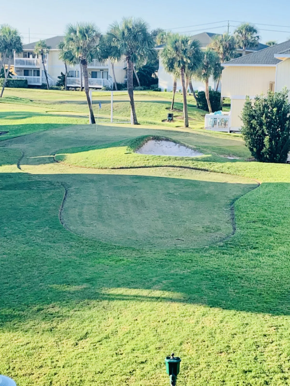 An exterior view of the putting green with a sand bunker, palm trees, and resort buildings with balconies in the background. The scene is calm, inviting, and recreational, perfect for enjoying a golf game in a coastal setting.