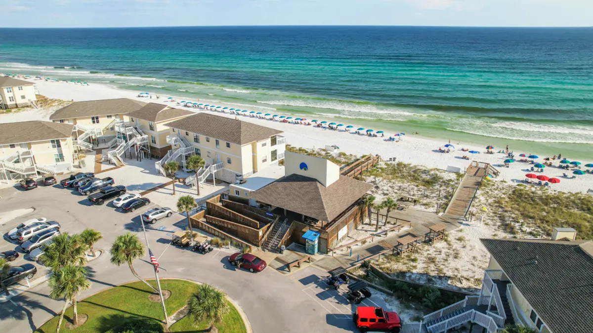 A stunning view of a white sandy beach dotted with colorful umbrellas, resort buildings, a boardwalk leading to the shore, and turquoise ocean waves. The scene is lively and inviting, evoking memories of sun-soaked days and easy access to the beach for family adventures.
