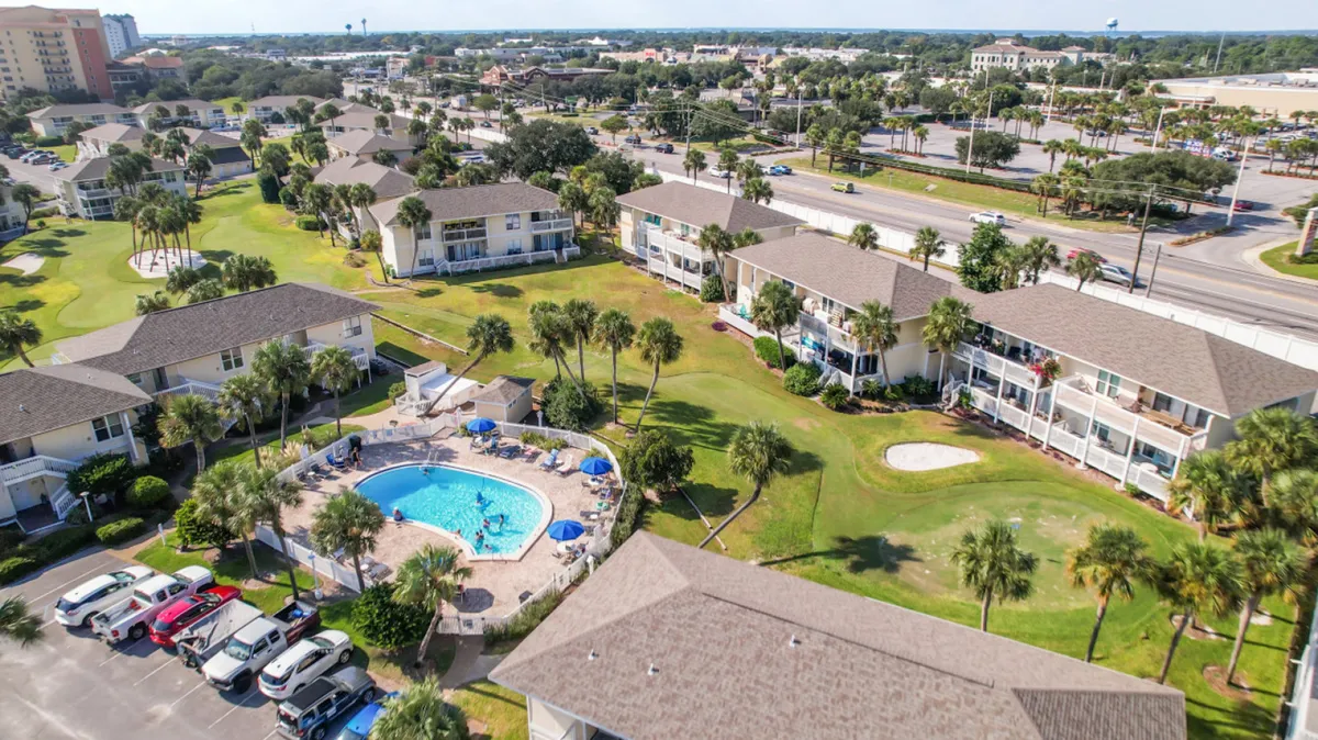 A central swimming pool surrounded by low-rise condo buildings with balconies, lush lawns, palm trees, and a nearby parking lot. People are enjoying the pool, and the easy access to both the condo and parking is evident.