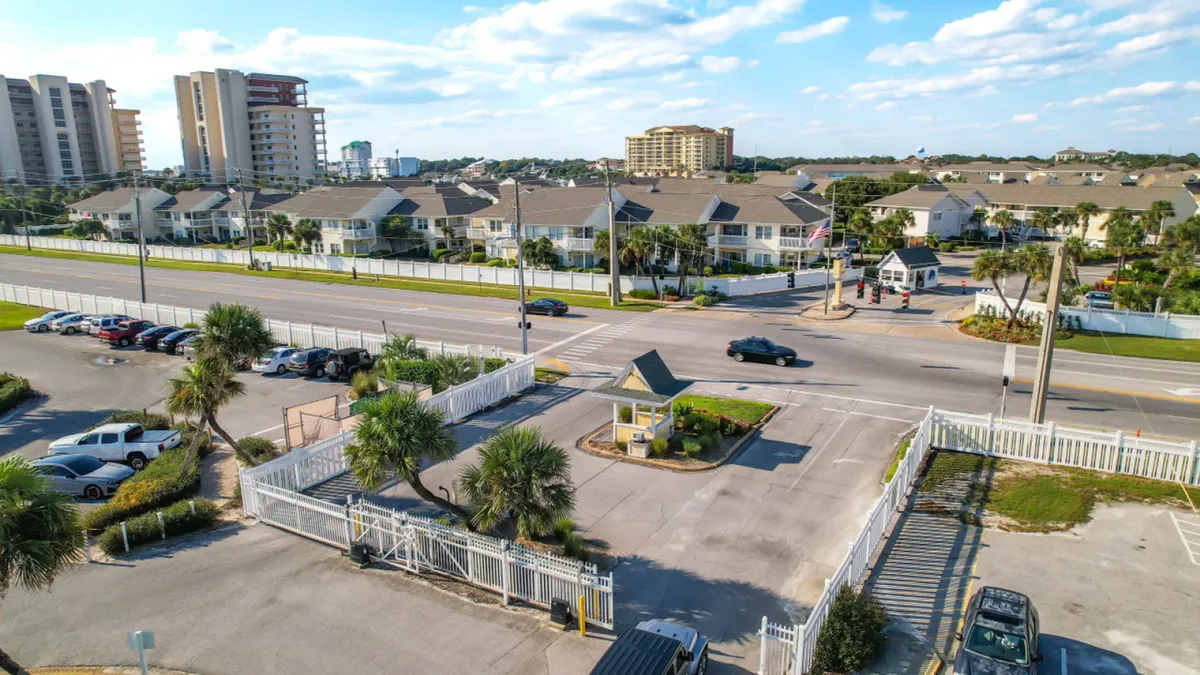 This image features the gated entrance to the resort, complete with a security booth, white fencing, and a well-maintained roadway. Residential buildings are visible in the background, reinforcing the sense of a protected, organized community.
