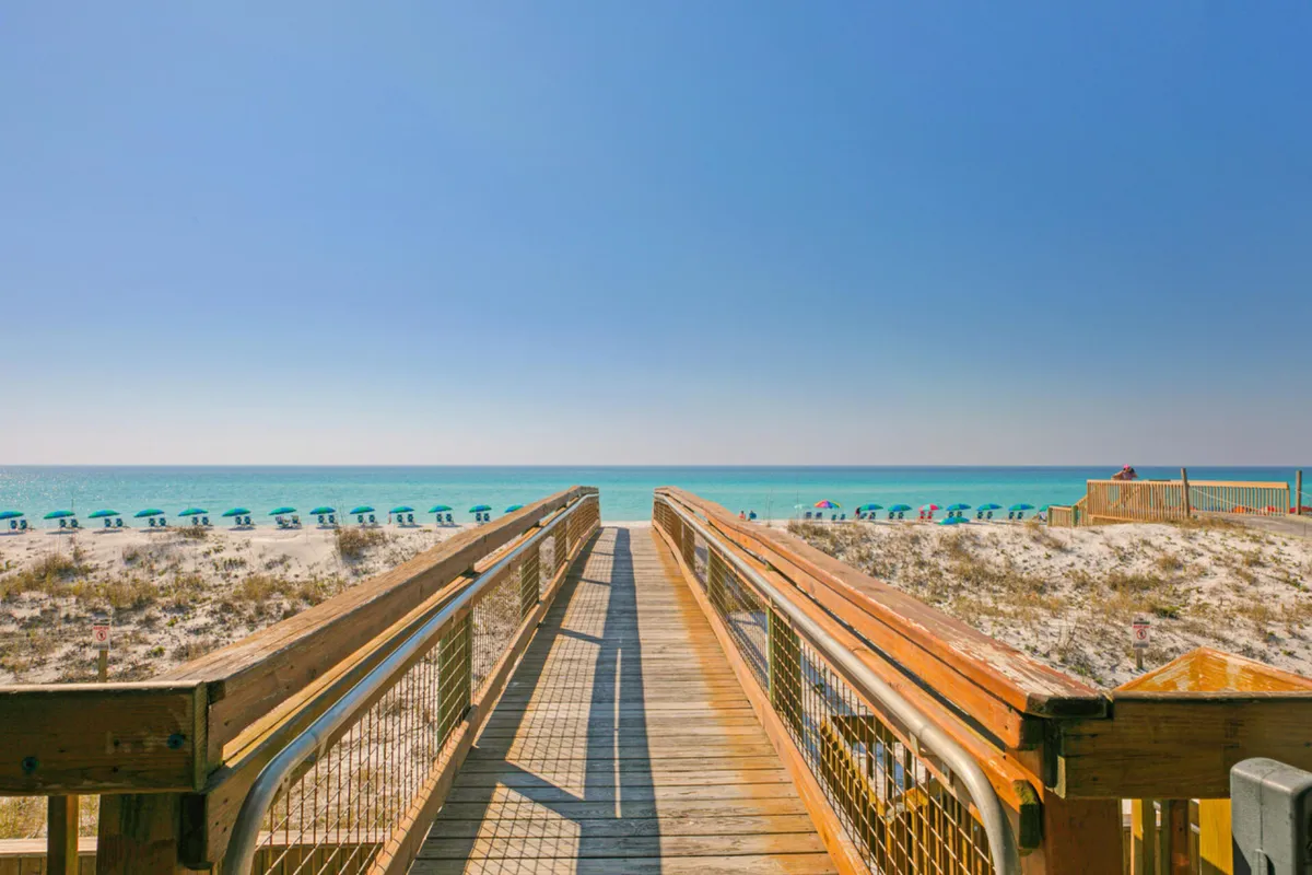 A wooden boardwalk leads directly to the turquoise ocean, flanked by soft white dunes and sparse coastal vegetation. A row of blue umbrellas lines the beach, inviting guests to relax under their shade. The expansive ocean horizon evokes anticipation for a day by the water, while a few people can be seen enjoying sunbathing and beach activities.