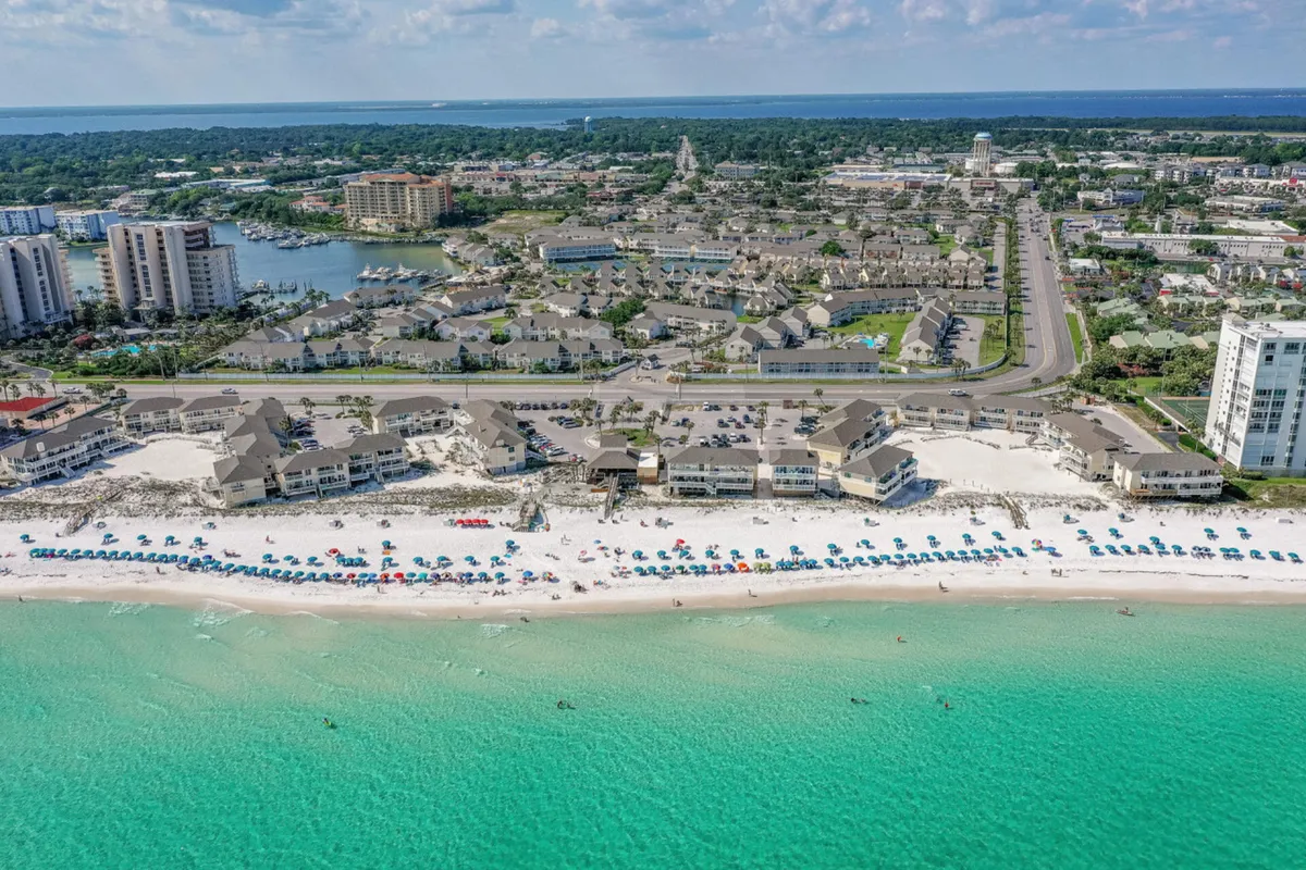 This image captures a pristine stretch of white sandy beach lined with organized rows of colorful beach umbrellas and lounge chairs, with the turquoise Gulf waters dotted with swimmers and beachgoers. In the background are beachfront condos and resort buildings, with a road and greenery just beyond, providing context for the proximity of accommodations to the beach.