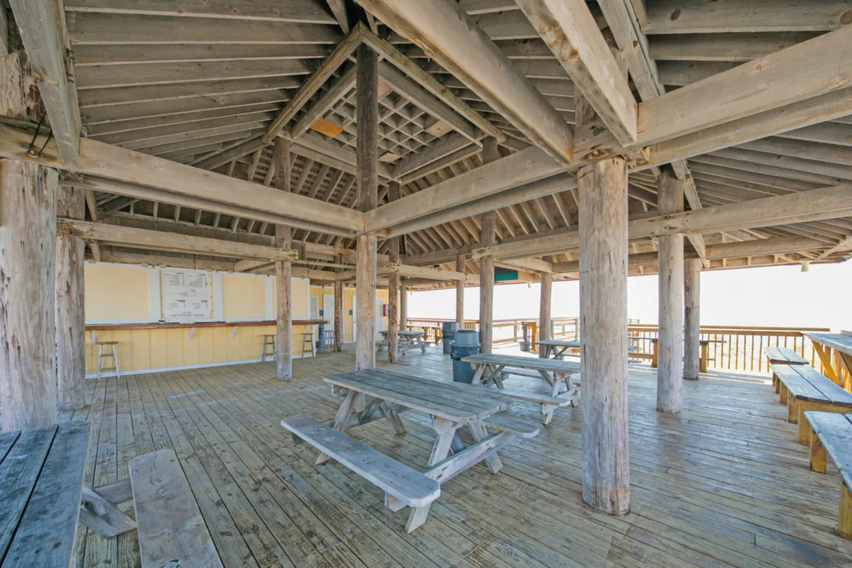 This image features a covered wooden deck with picnic tables, open sides with ocean views, and a relaxed, communal atmosphere. Diners can enjoy the fresh sea air while gathering for meals or drinks, with a scenic harbor visible in the background.